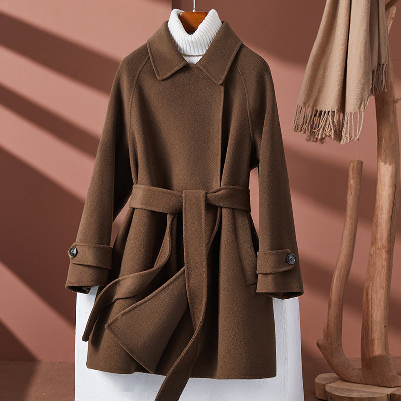 A brown coat with a belt, displayed on a mannequin set against a neutral background. The coat features a flared silhouette and is paired with a white sweater.